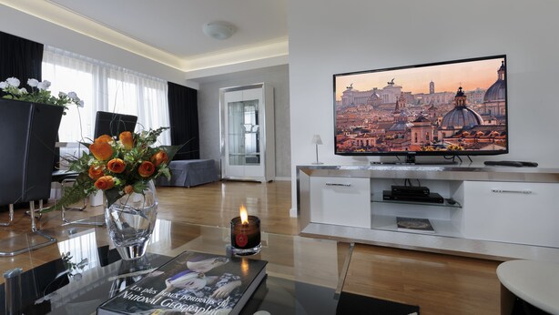 Una sala de estar moderna con una televisión de pantalla plana mostrando un paisaje urbano, un florero con flores y un libro sobre una mesa de vidrio.