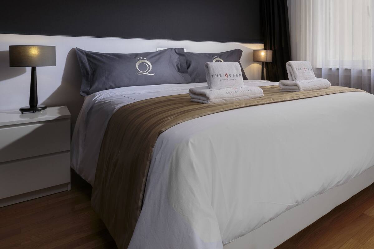 A neatly made bed with a white comforter and a brown throw blanket, two pillows, a bedside table with a lamp, and a window with sheer curtains.