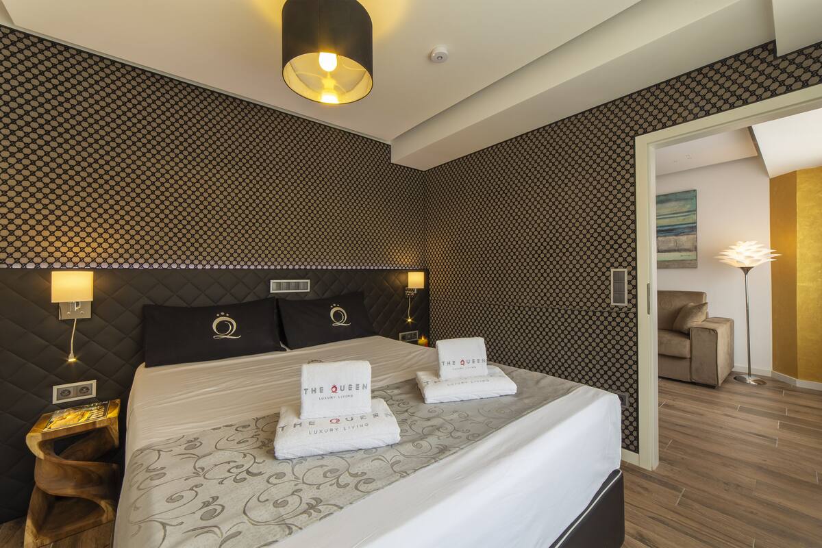A hotel room with a large bed, two pillows with a logo, a bedside table with a lamp, and a patterned wall.