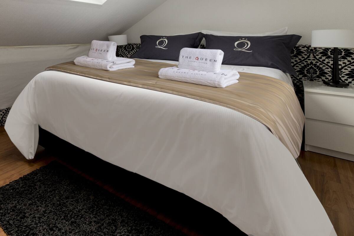 A neatly made bed with a white comforter and two folded towels with "The Queen" branding, a black and white patterned bench, and a small table with a lamp.