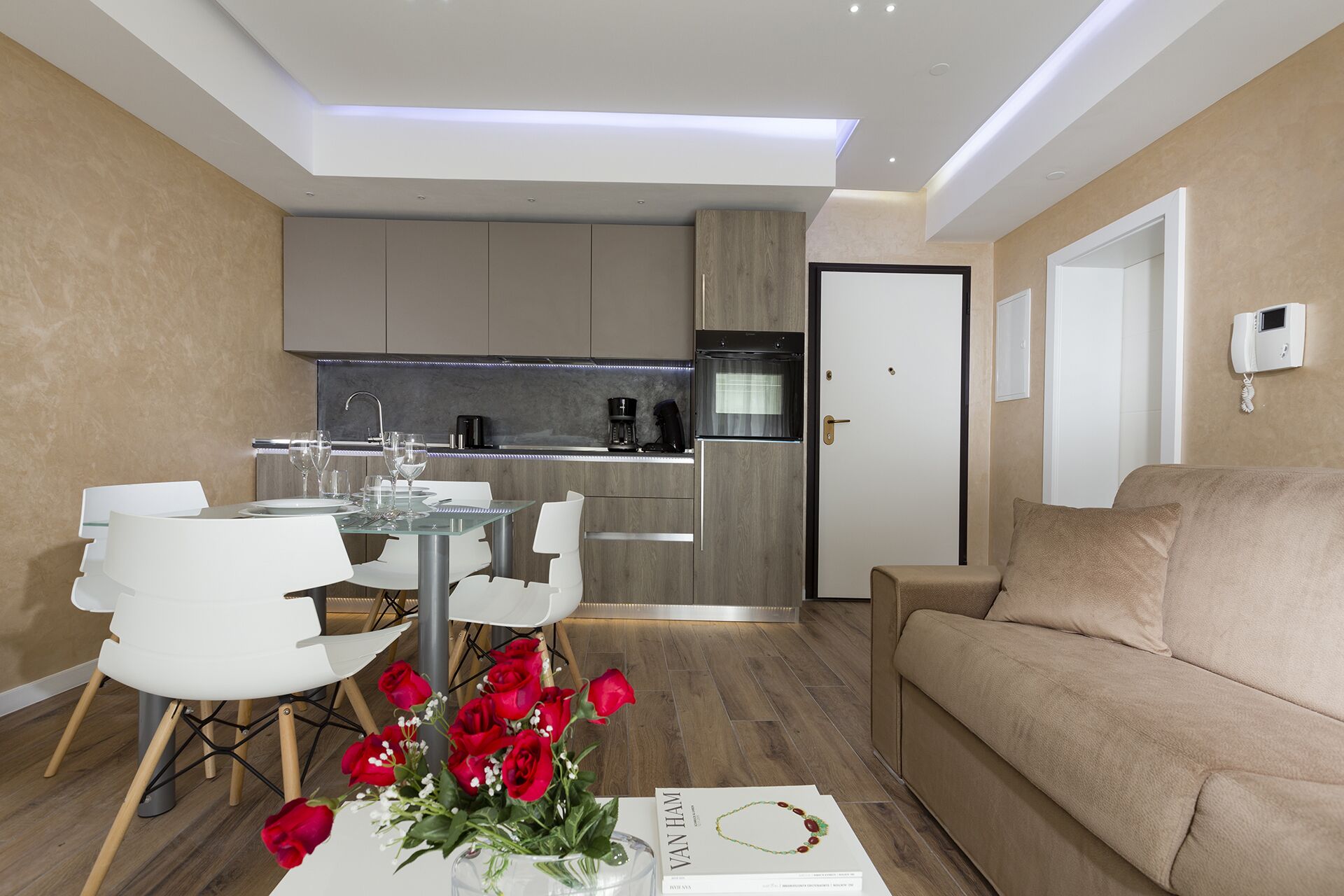 Een moderne woonkamer met een beige bank, een glazen eettafel met witte stoelen, en een keuken met grijze kastjes en een inbouwoven.