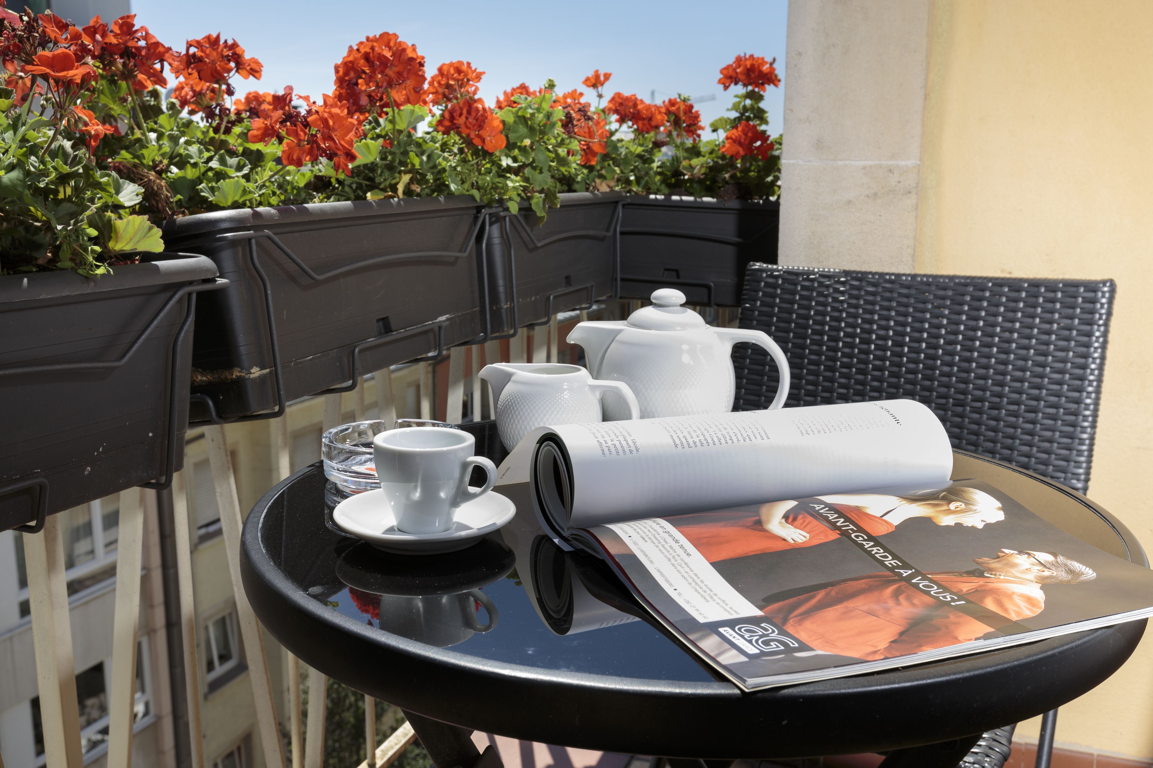 A table with a magazine, a teapot, a cup, and saucer on a balcony with red flowers in the background.