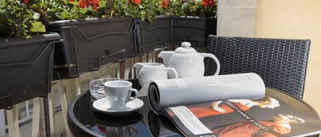 A table with a magazine, a teapot, a cup, and saucer on a balcony with red flowers in the background.