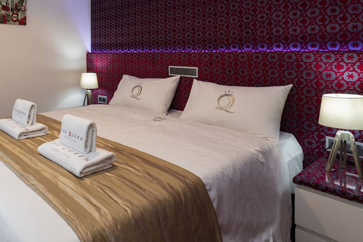 A hotel room with a large bed, two bedside tables with lamps, a mirror, and a wall with a red patterned wallpaper.