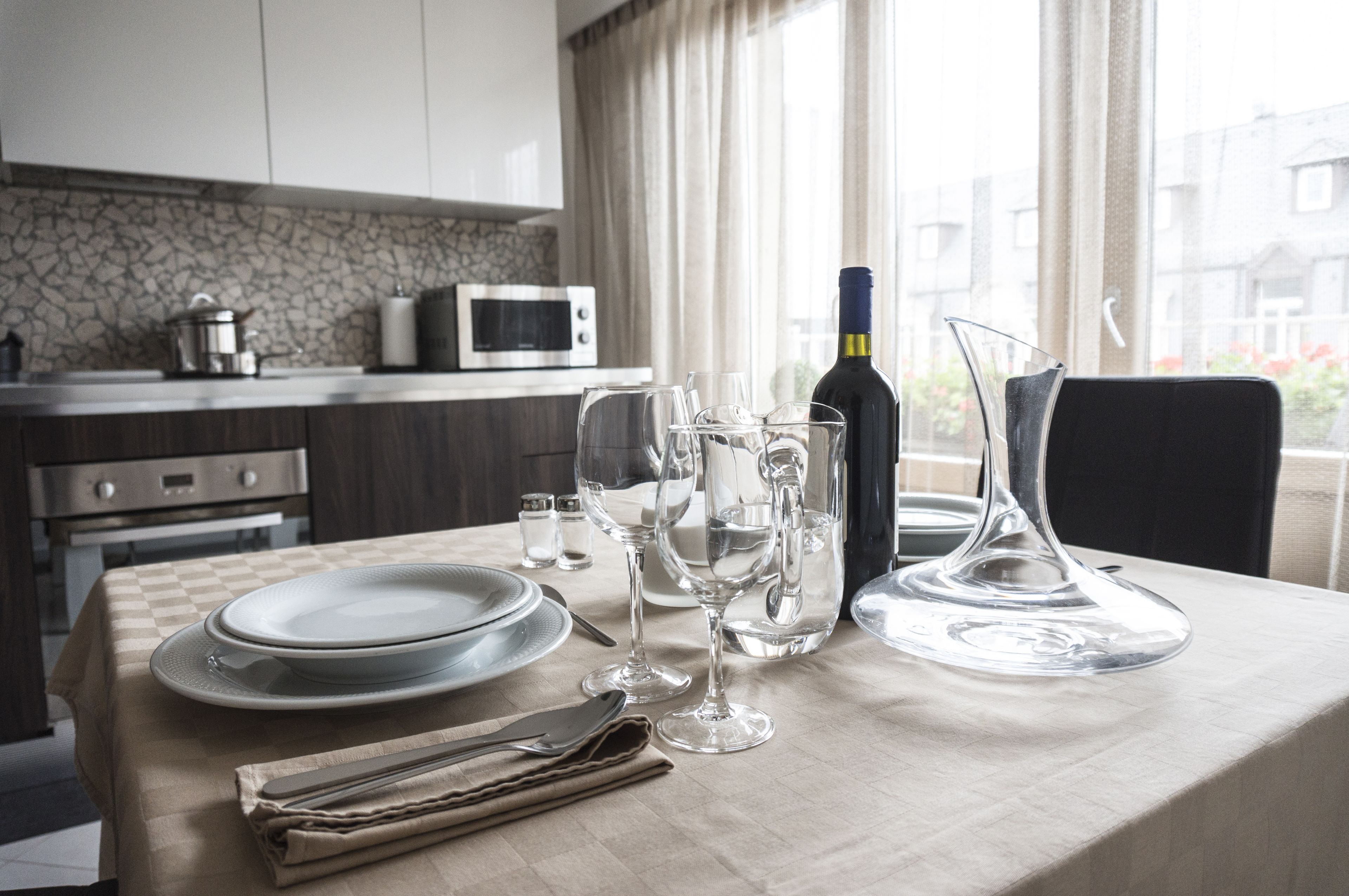 A neatly set dining table with plates, glasses, a wine bottle, and a pitcher in a modern kitchen with a stone backsplash and a view of buildings outside.
