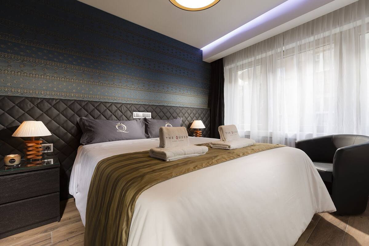 A modern hotel room with a large bed, a dark patterned headboard, and a large window with sheer curtains.