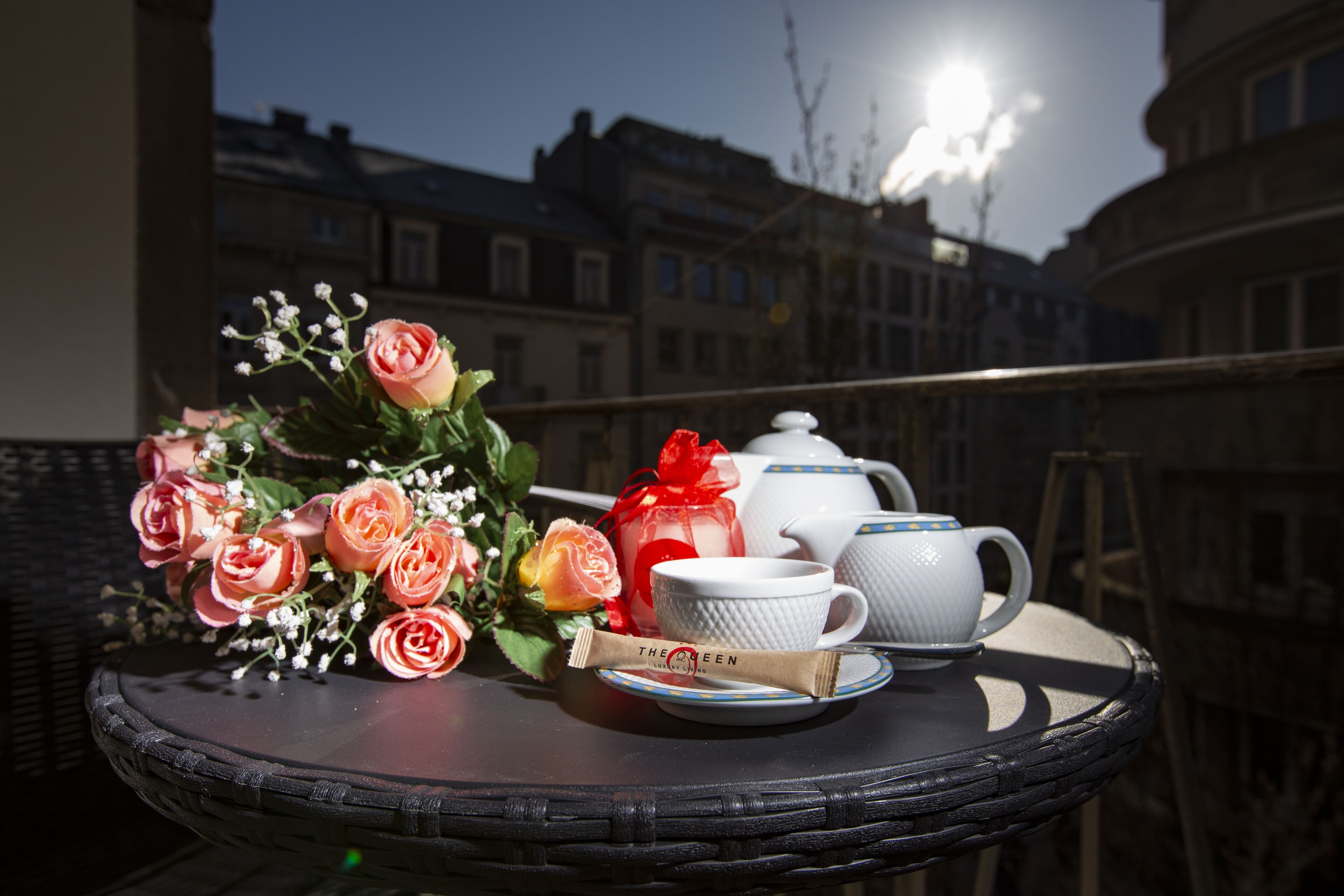Uma mesa de vime com um conjunto de chá e um buquê de rosas numa varanda com vista para edifícios.