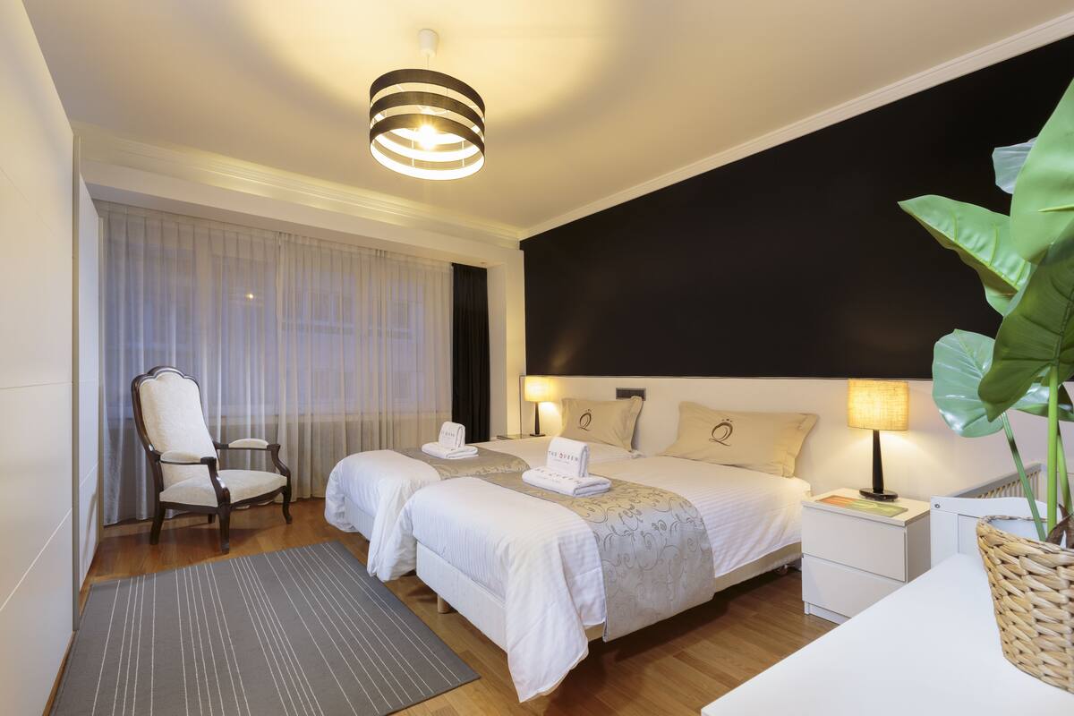 A hotel room with two beds, a chair, a bedside table with a lamp, a large plant, and a view of the outside through sheer curtains.