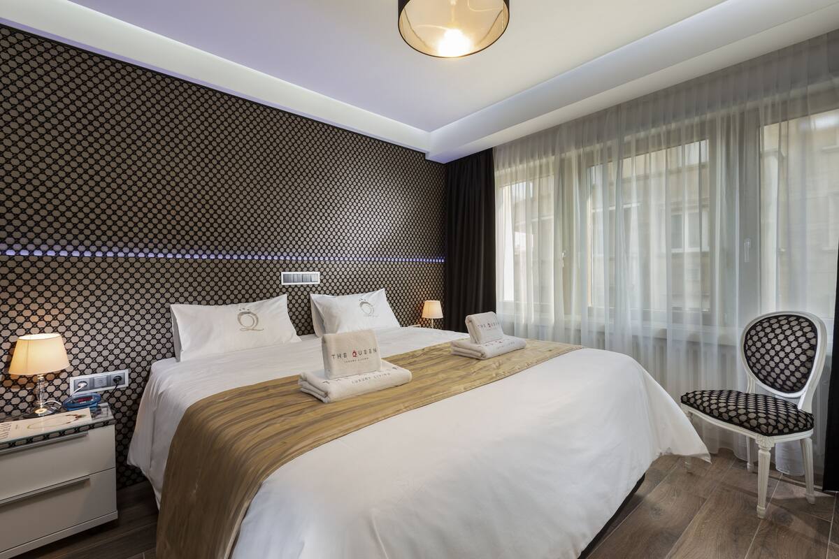 A modern hotel room with a large bed, a nightstand with a lamp, a chair, and a patterned wall.