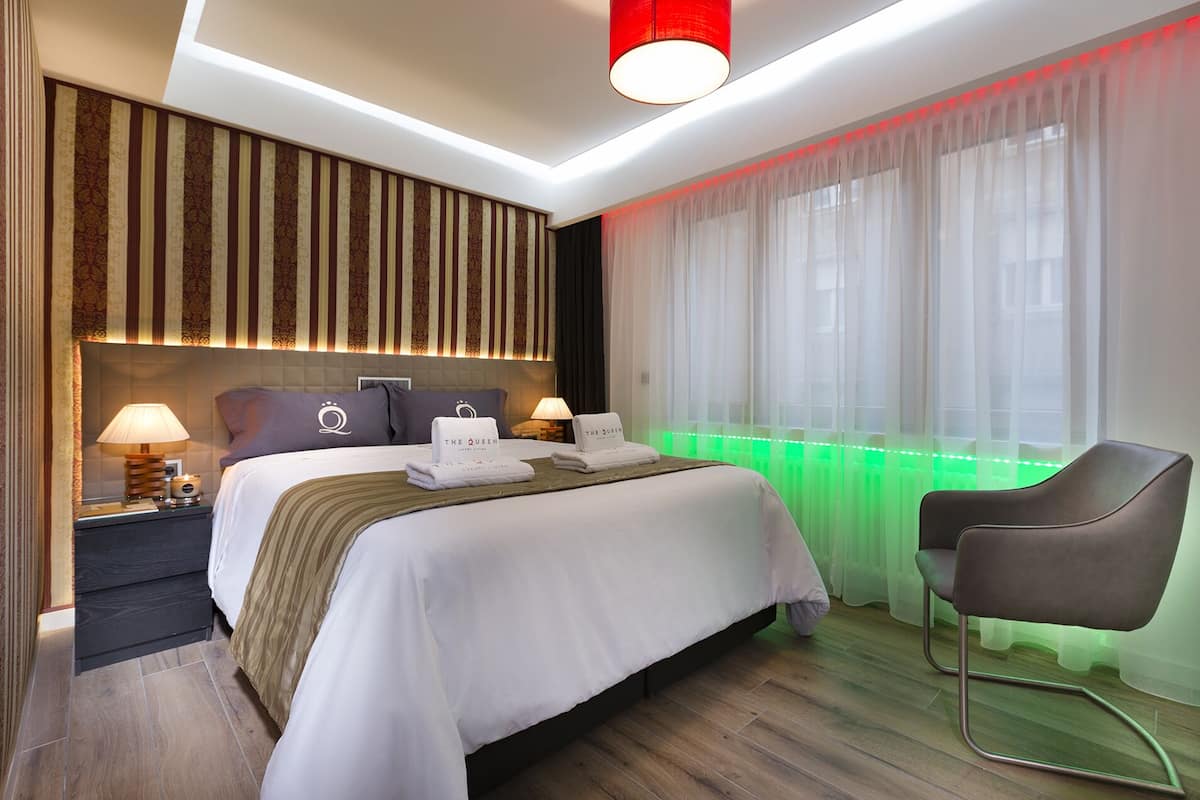 A modern hotel room with a large bed, two bedside tables, a desk, a chair, and a window with sheer curtains and green LED lighting.