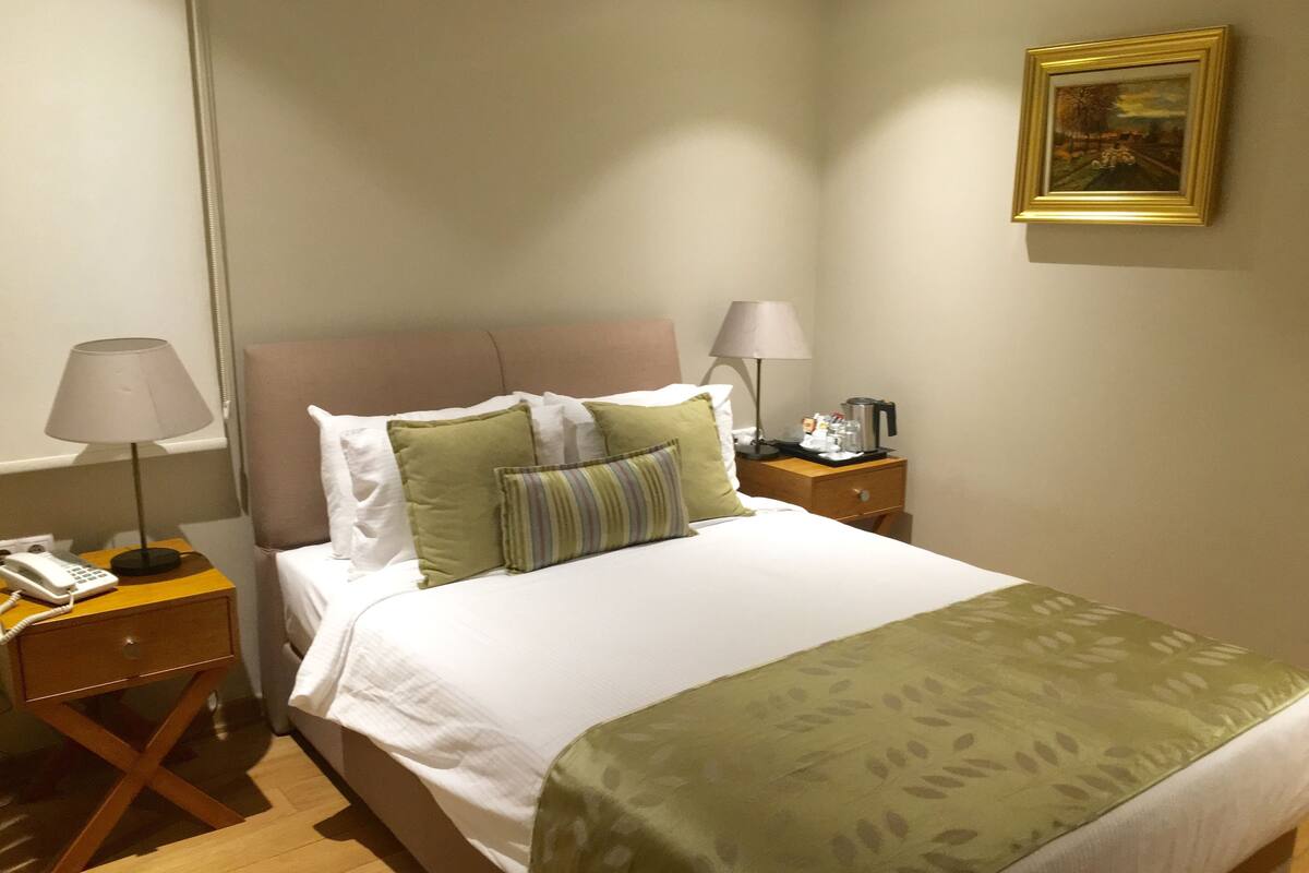 Una cama bien hecha con una manta verde, dos lámparas de noche, una mesita con un teléfono y un cuadro en la pared.