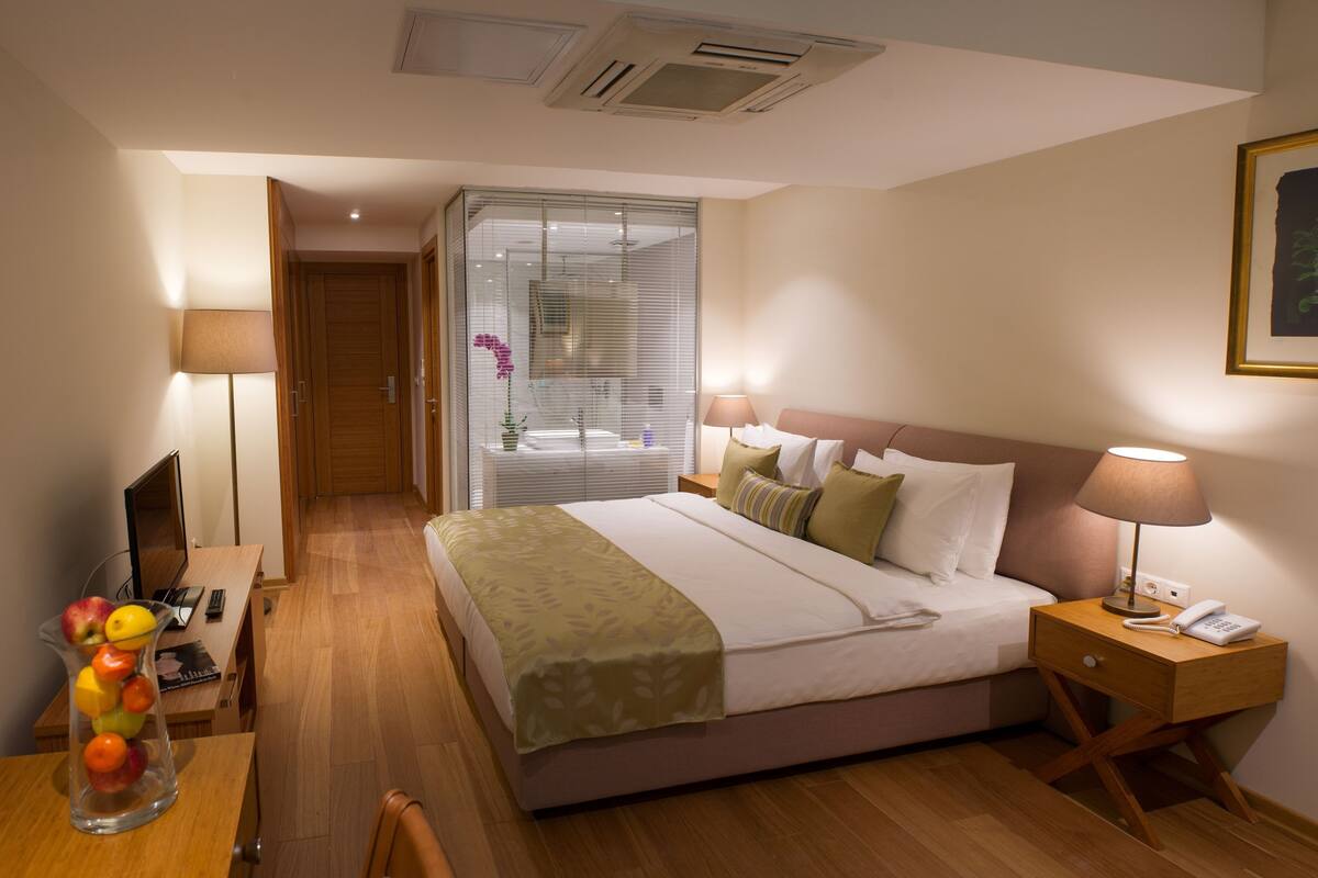 Una habitación de hotel con una cama, lámparas de noche, un escritorio con una computadora, un florero con fruta y una ventana con persianas.