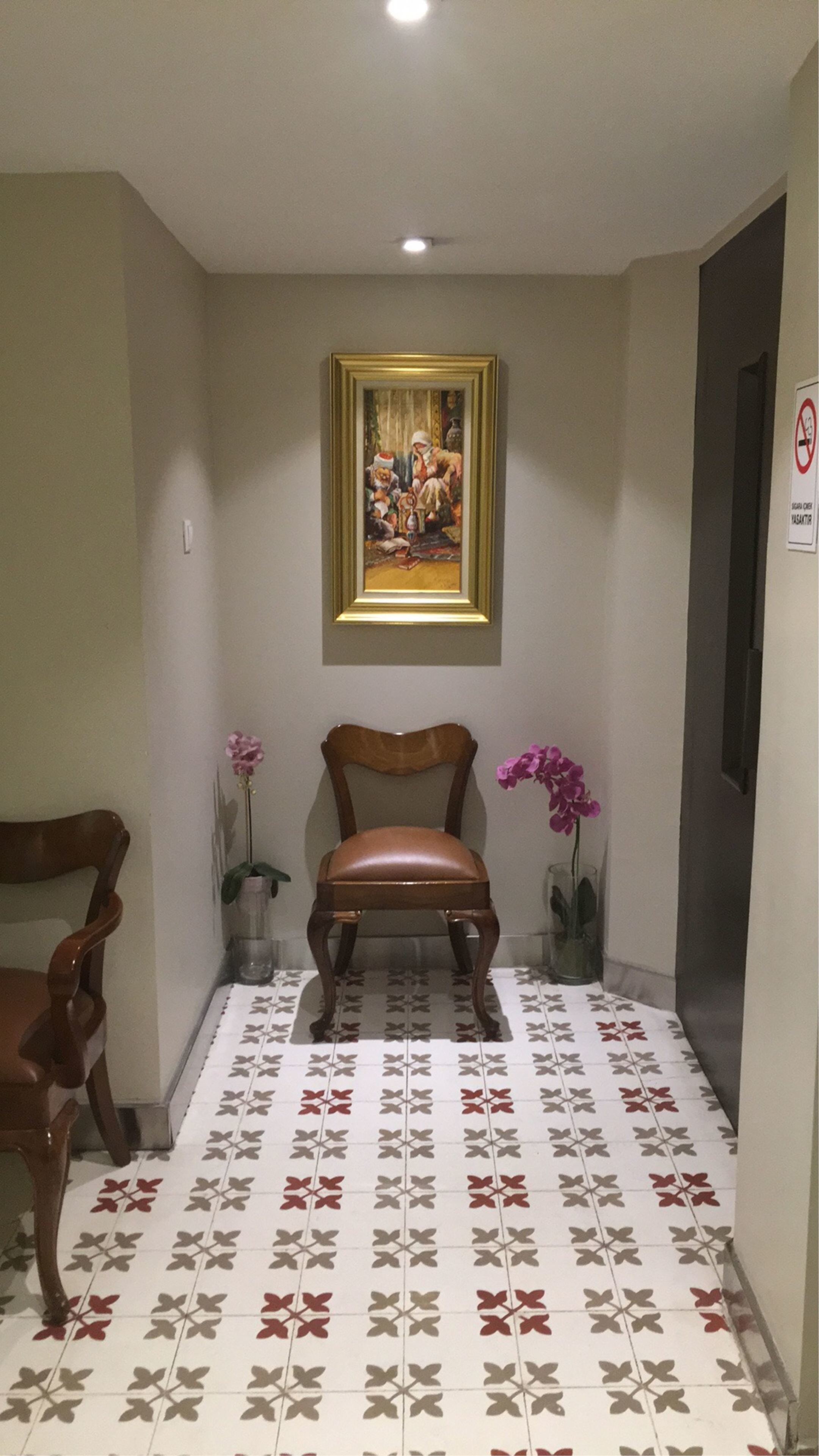 A hallway with a patterned floor, a framed painting on the wall, a wooden chair, and two potted plants.