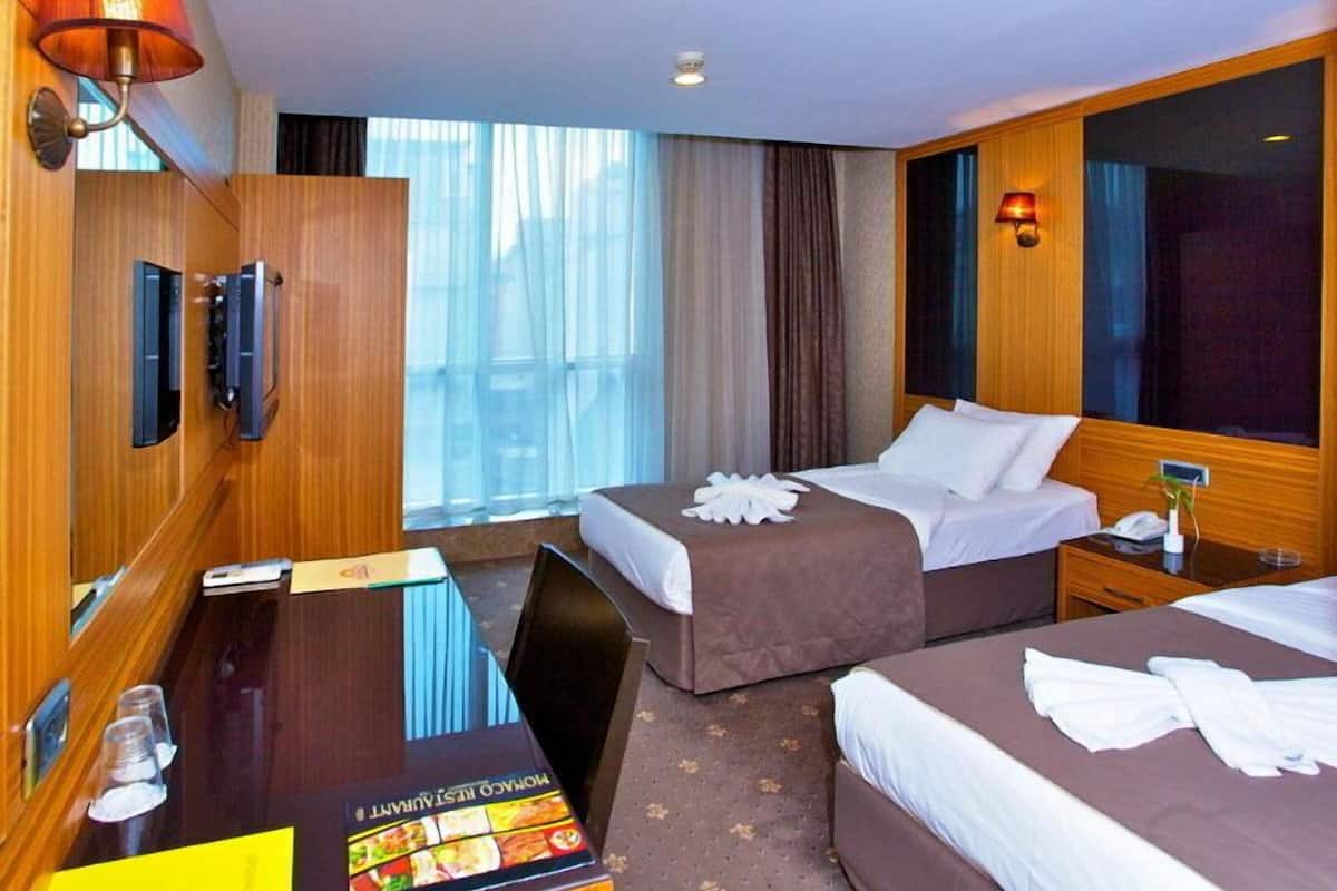 A hotel room with two beds, a desk, a minibar, and a large window with curtains.