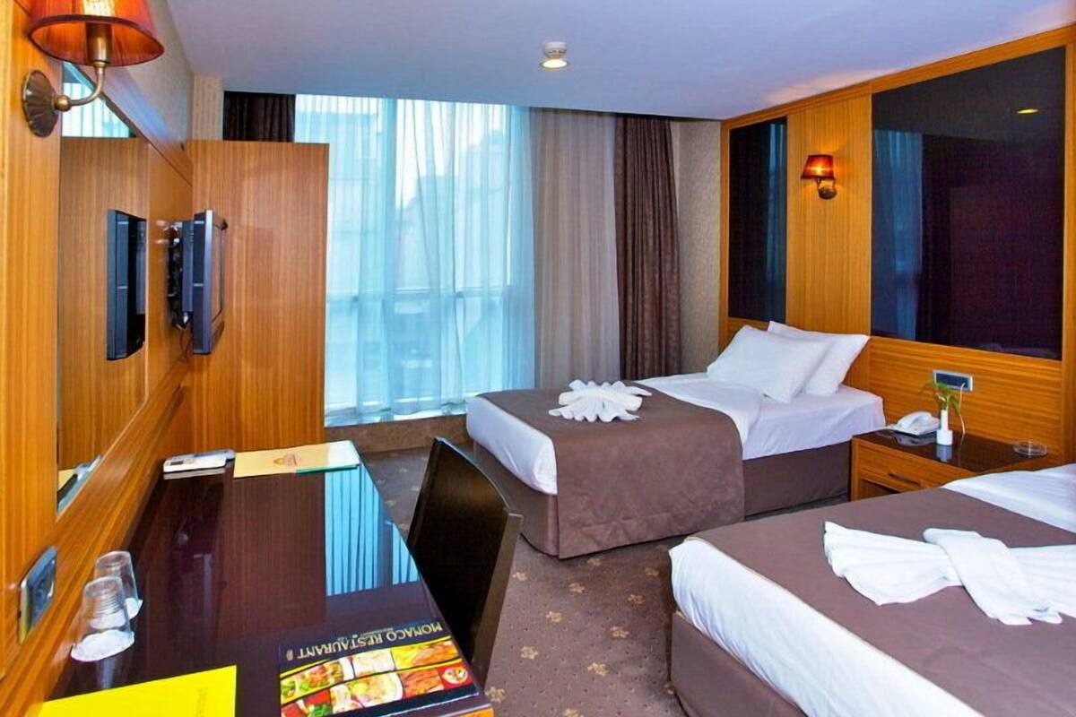 A hotel room with two beds, a desk, a minibar, and a large window with curtains.