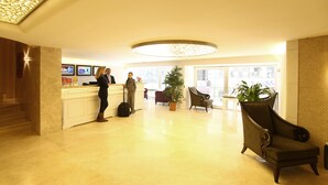 Un vestíbulo de hotel con una recepción, una planta grande en maceta, un área de estar con una silla y una escalera que sube a otro nivel.