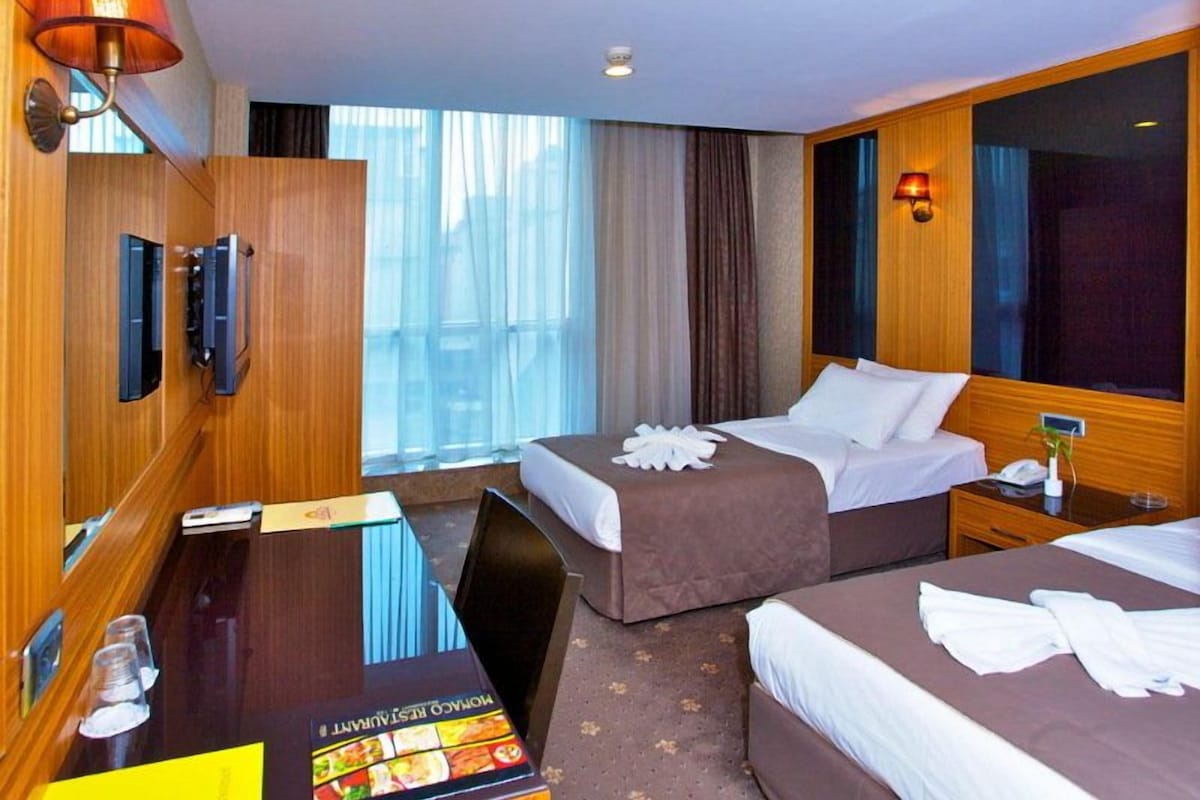 A hotel room with two beds, a desk, a minibar, and a large window with curtains.