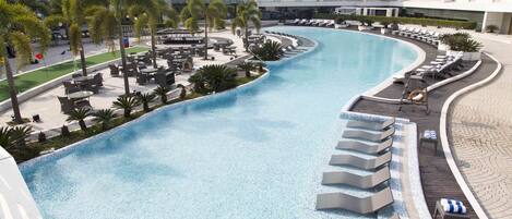 A curved swimming pool with lounge chairs and palm trees, surrounded by modern buildings.