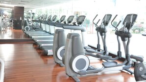A row of stationary bikes in a gym with large windows and a wooden floor. - The Deltin Hotel (Daman)