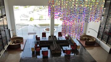 Um lobby moderno com piso de mármore refletivo, área de estar central com sofás e mesas, e uma grande janela com vista para a área externa.