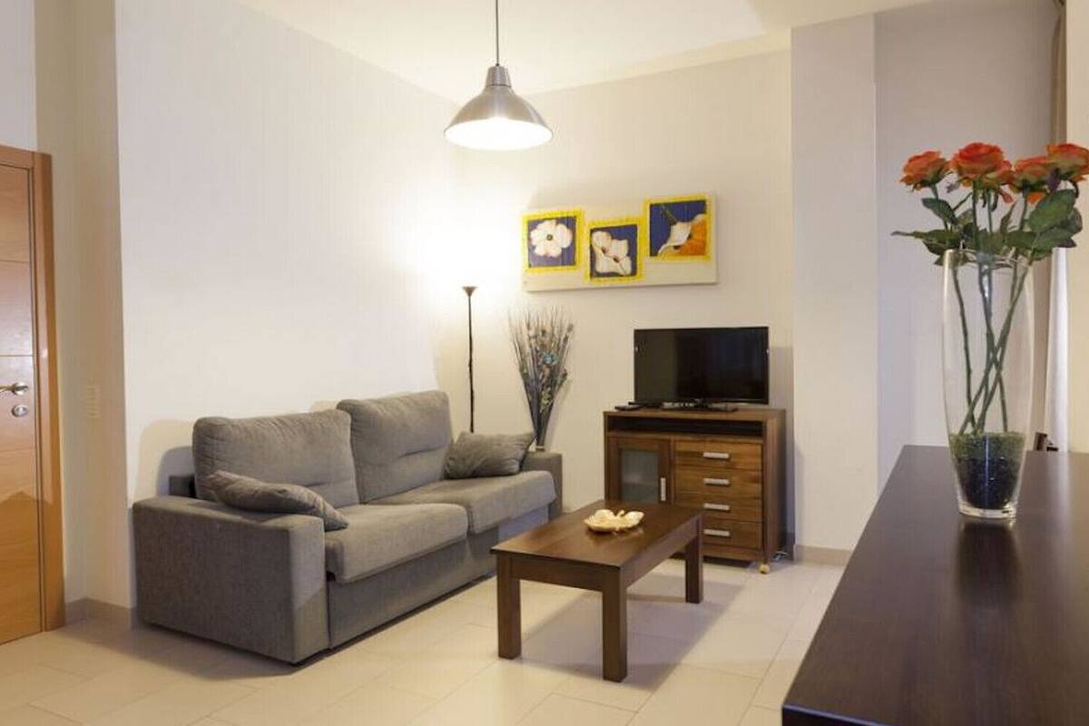 Una sala de estar moderna con un sofá gris, una mesa de centro de madera, un televisor sobre un mueble de madera y un jarrón con flores sobre una mesa.
