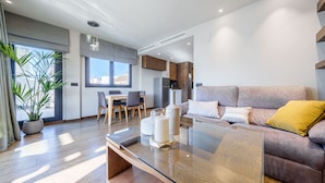 Una sala de estar moderna con una mesa de centro de vidrio, un sofá con cojines, una zona de comedor con mesa y sillas, y un ventanal con vistas a edificios.