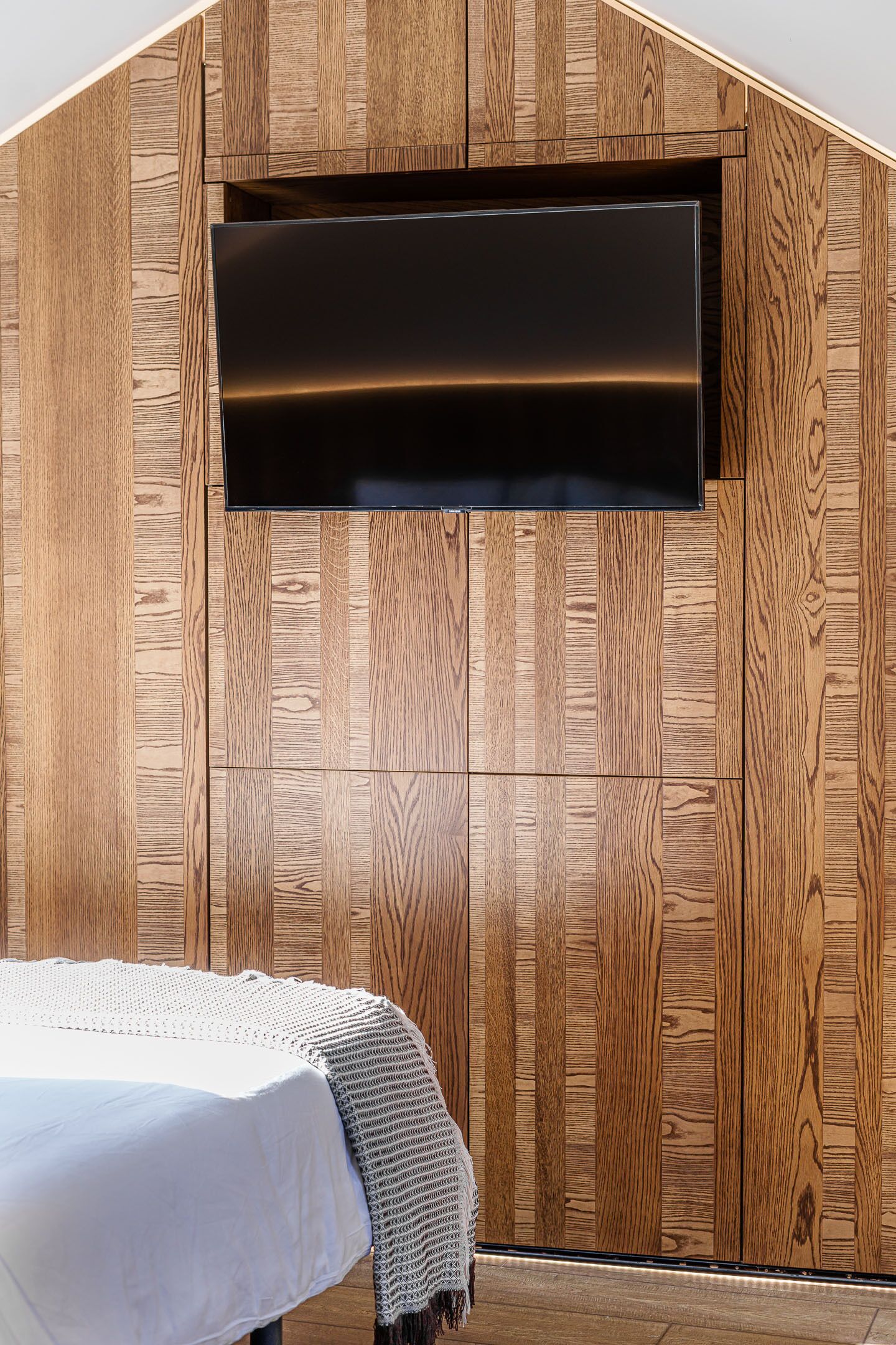 A room with a wooden wall panel design, a flat-screen TV mounted on the wall, and a bed with a white bedspread and a knitted throw.