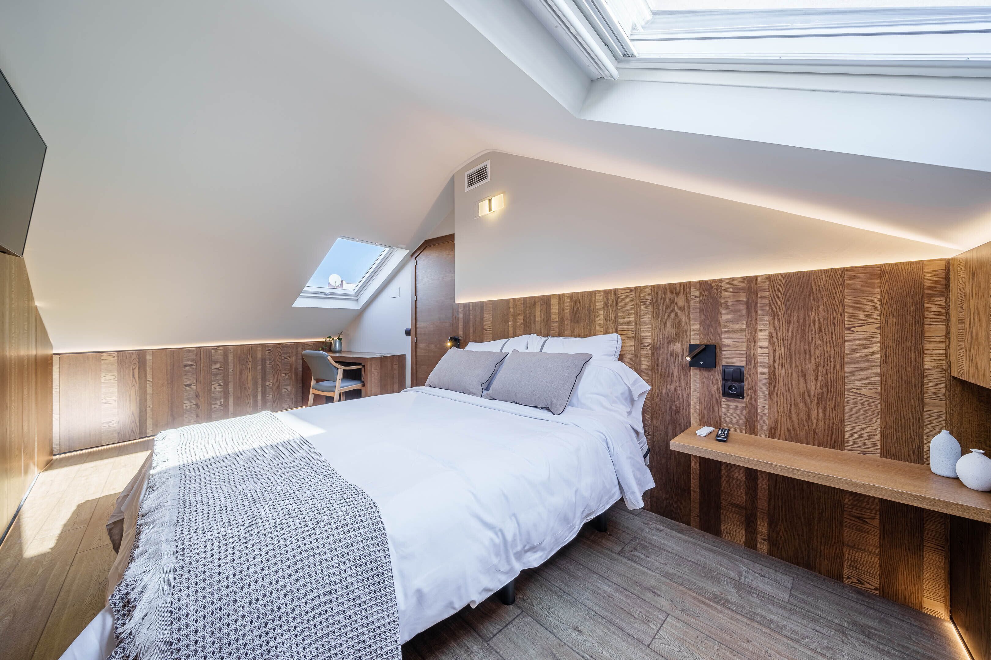 Ein modernes Schlafzimmer mit Oberlicht, Holzwandverkleidung, einem Bett mit weißer Bettwäsche, einem Nachttisch mit Lampe und einem kleinen Schreibtisch mit Stuhl.