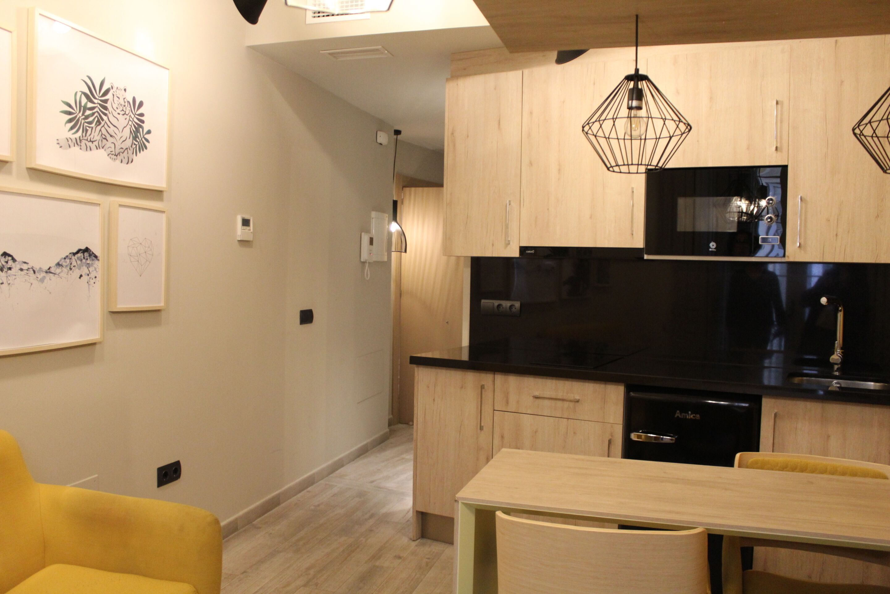 A modern kitchen with wooden cabinets, a black microwave, and a dishwasher. There is a dining table with chairs and a yellow armchair.