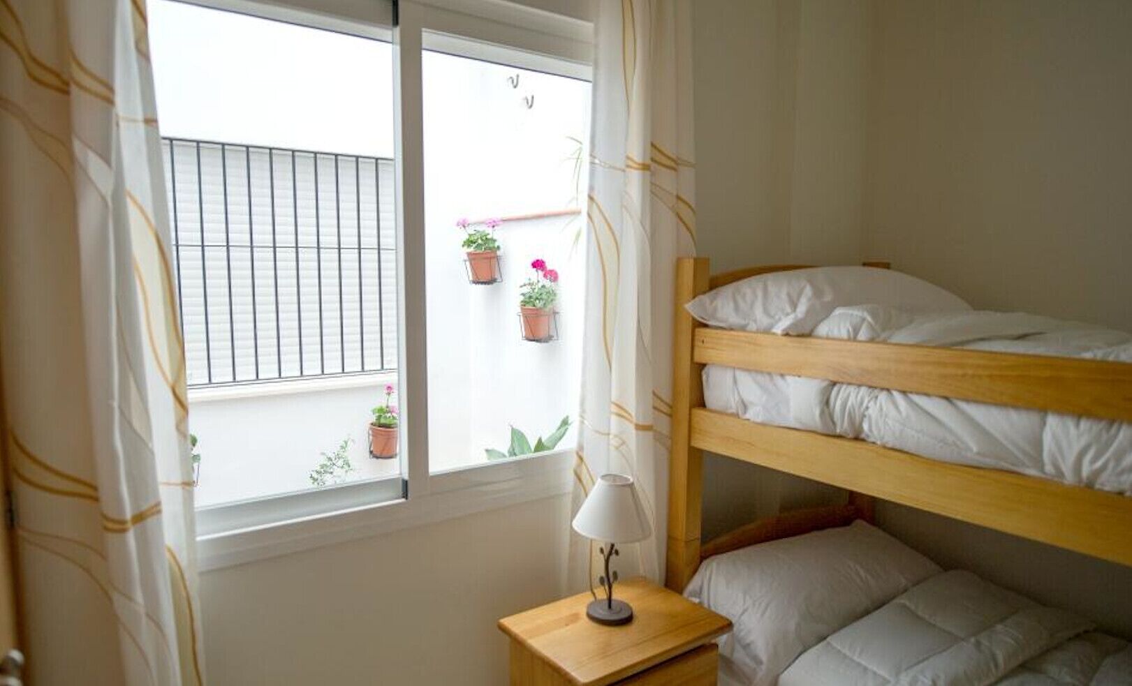 Une chambre avec un lit superposé, une fenêtre, une table de chevet avec une lampe et des plantes en pot sur le rebord de la fenêtre.