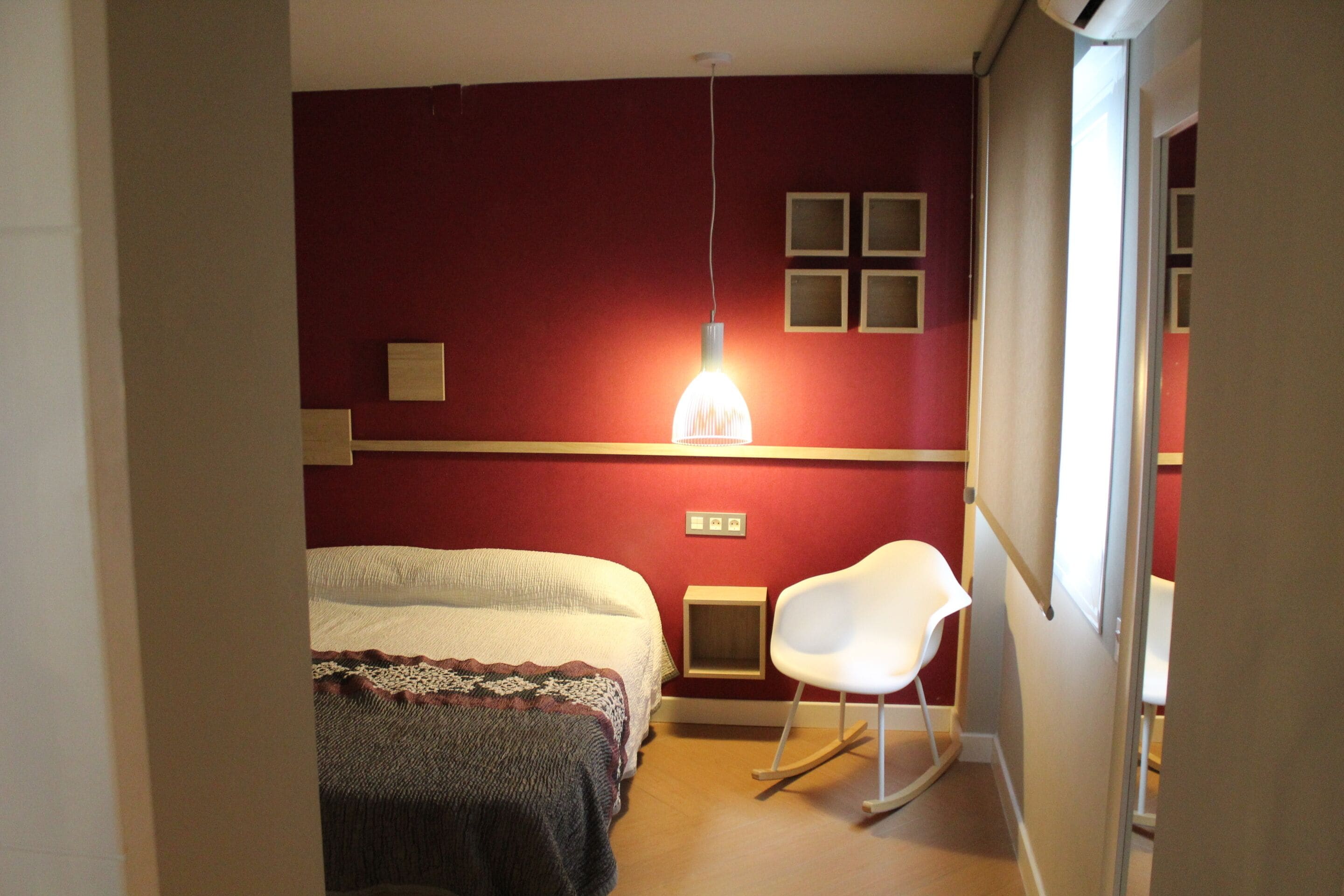 A bedroom with a bed, a white rocking chair, a hanging light fixture, and a red wall.
