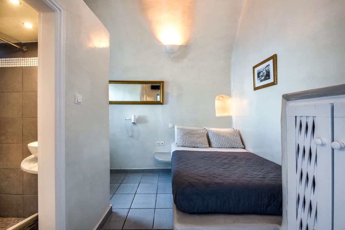 A compact hotel room with a bed, a bedside table, a mirror, a wall-mounted light, a toilet, and a shower area.