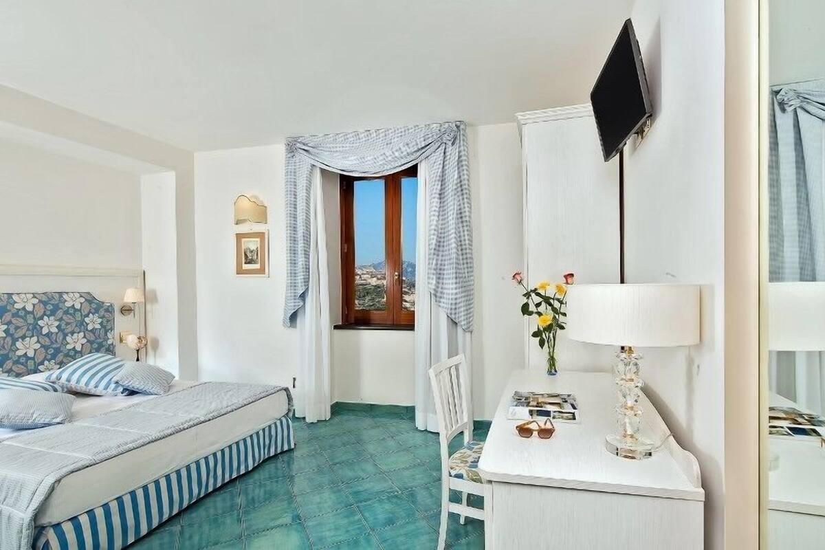 Habitación de hotel con una cama, un escritorio con lámpara, un televisor montado en la pared y una ventana con cortinas.