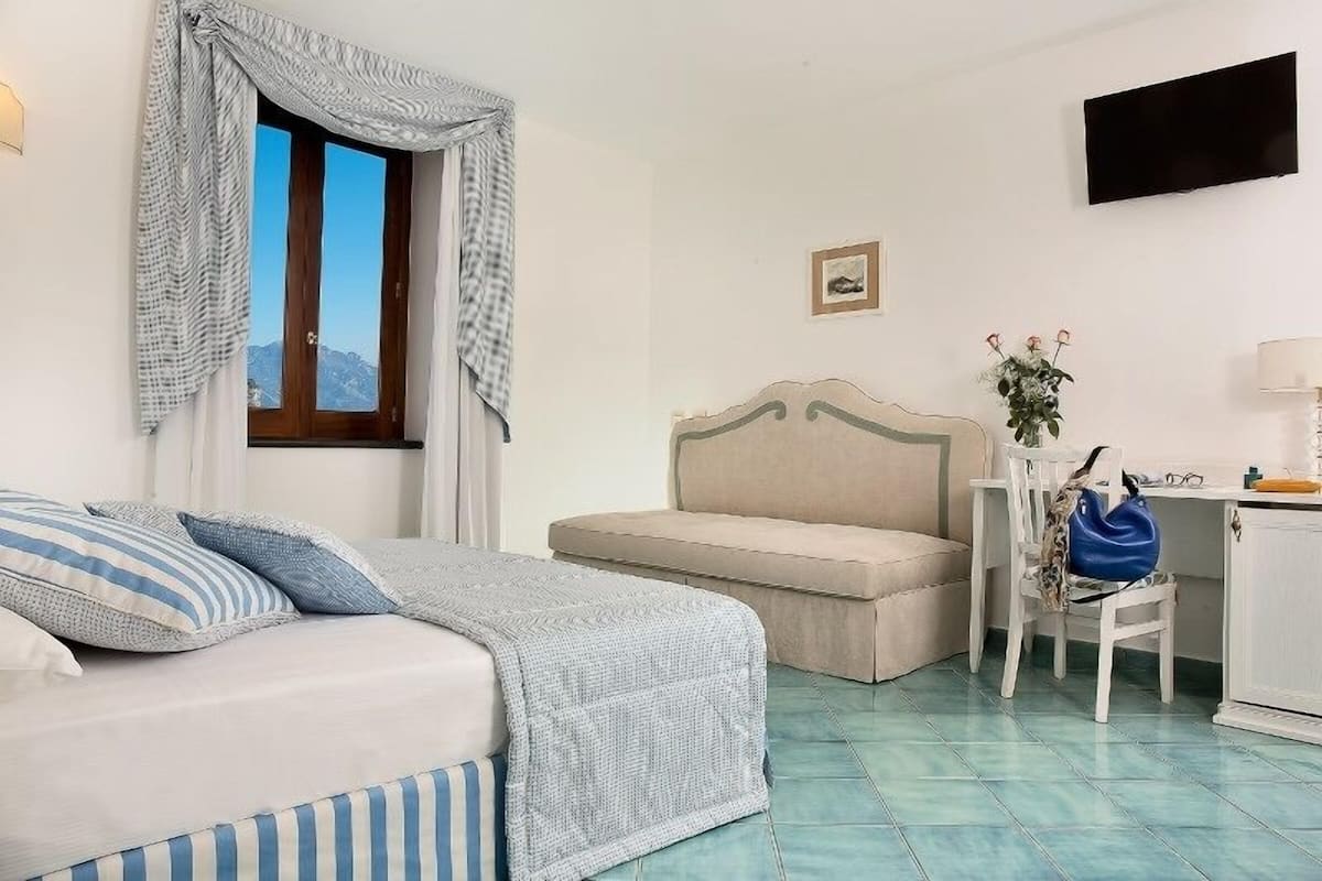 Un dormitorio con cama, un banco, un escritorio, una silla, un televisor y una ventana con cortinas.