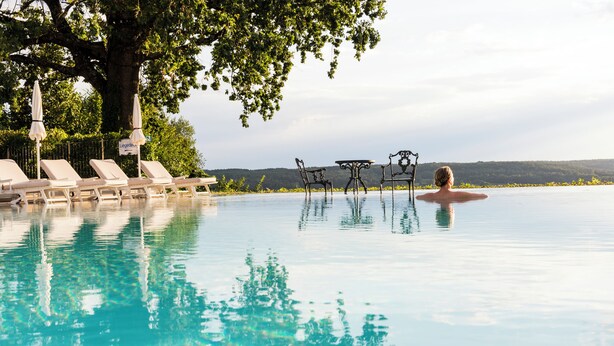 A pool with lounge chairs and a table, surrounded by trees and a view of the landscape. - Hotel & Spa Der Steirerhof Bad Waltersdorf (Bad Waltersdorf)