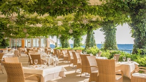 An outdoor dining area with wicker chairs and tables, covered by a leafy canopy, overlooking a scenic view. - Hotel & Spa Der Steirerhof Bad Waltersdorf (Bad Waltersdorf)