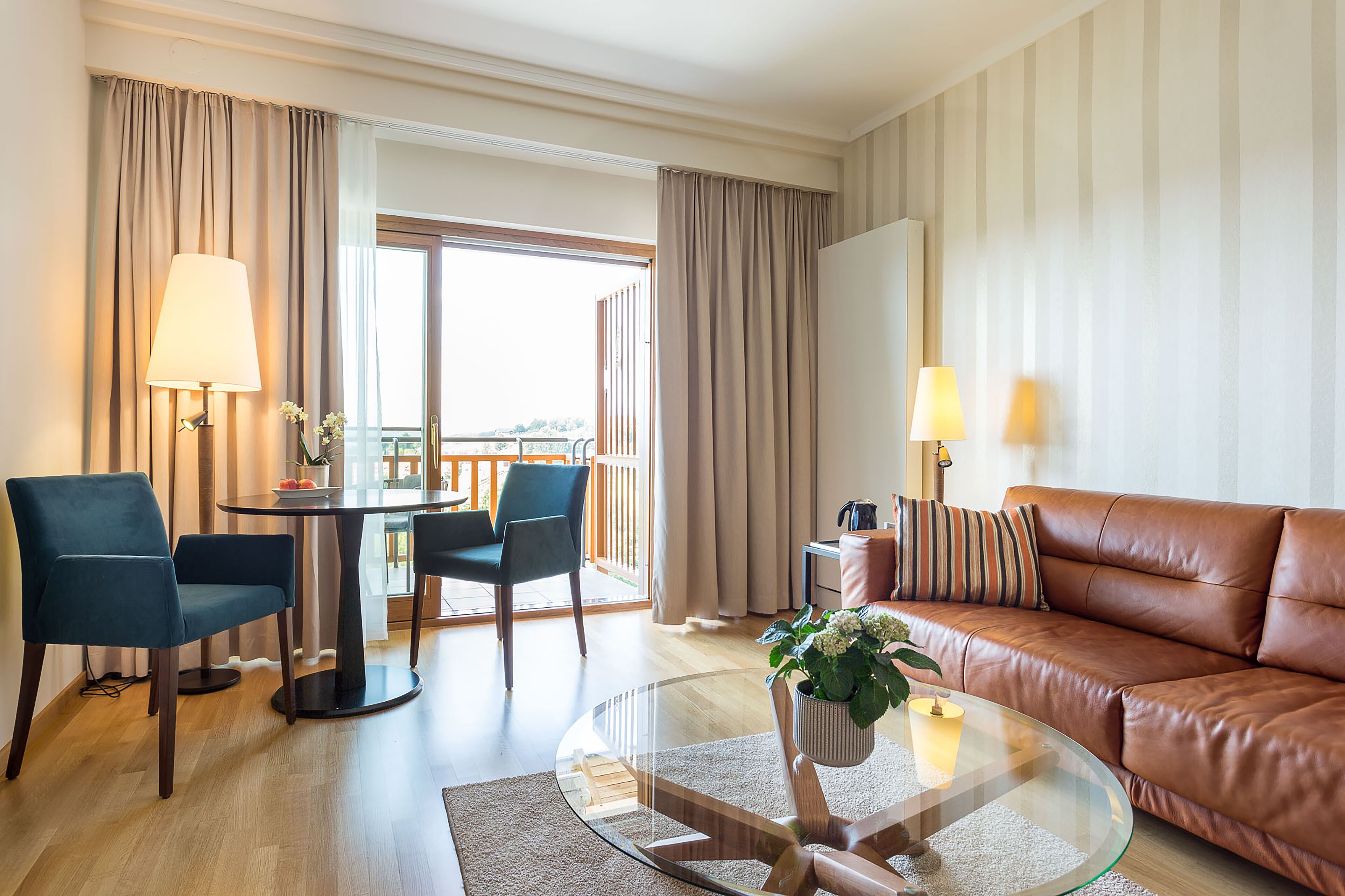 A hotel room with a glass coffee table, a leather sofa, two armchairs, a dining table with chairs, and a balcony with curtains.