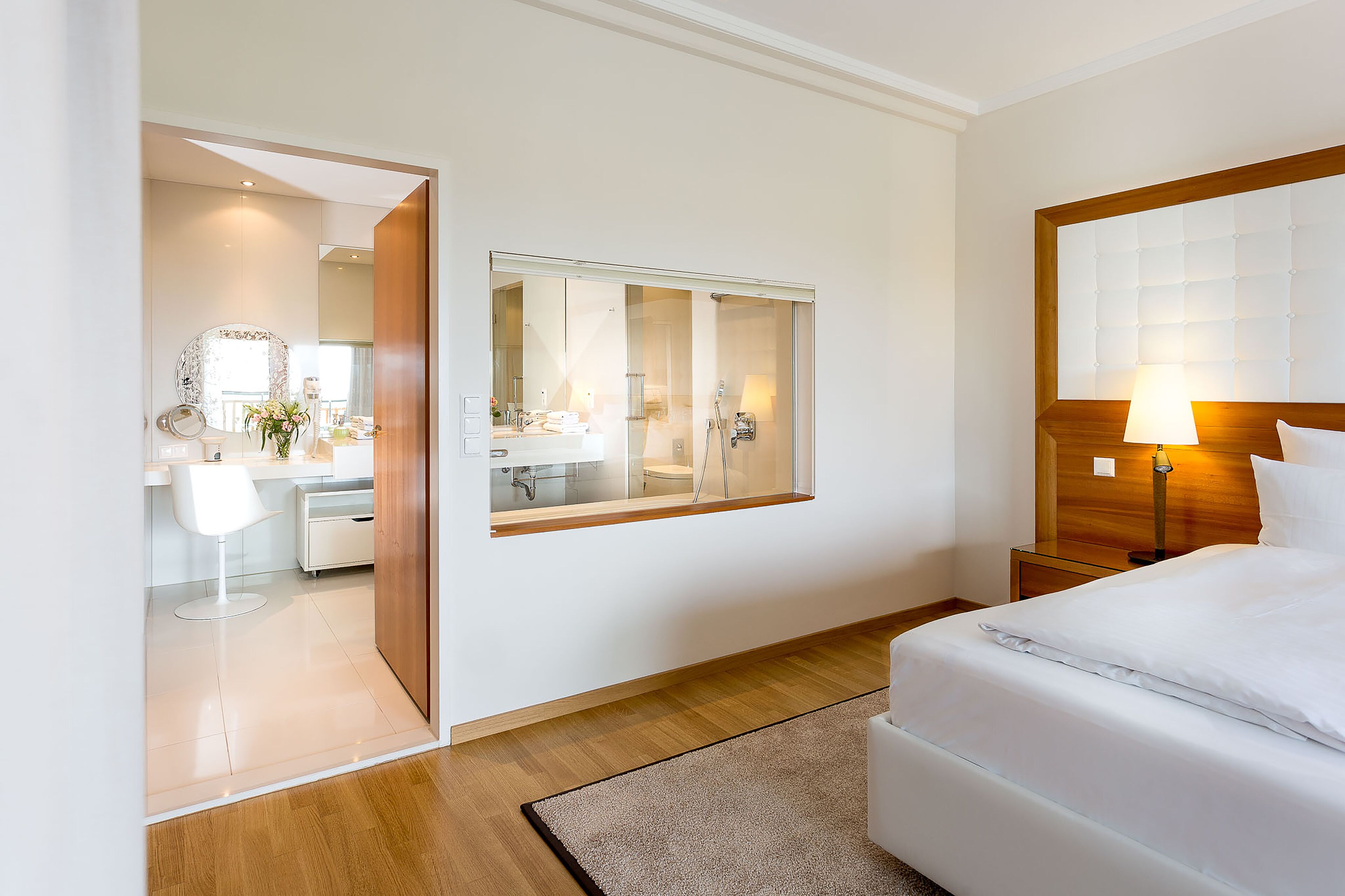 A modern hotel room with a large bed, a bedside table with a lamp, a mirror, and a view of the bathroom through a glass door.