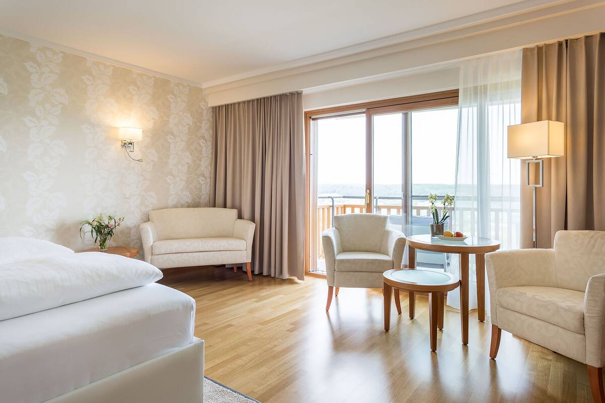 Ein Hotelzimmer mit einem großen Bett, zwei Sesseln, einem kleinen Tisch mit einer Lampe und Blick auf die Stadt durch große Fenster.