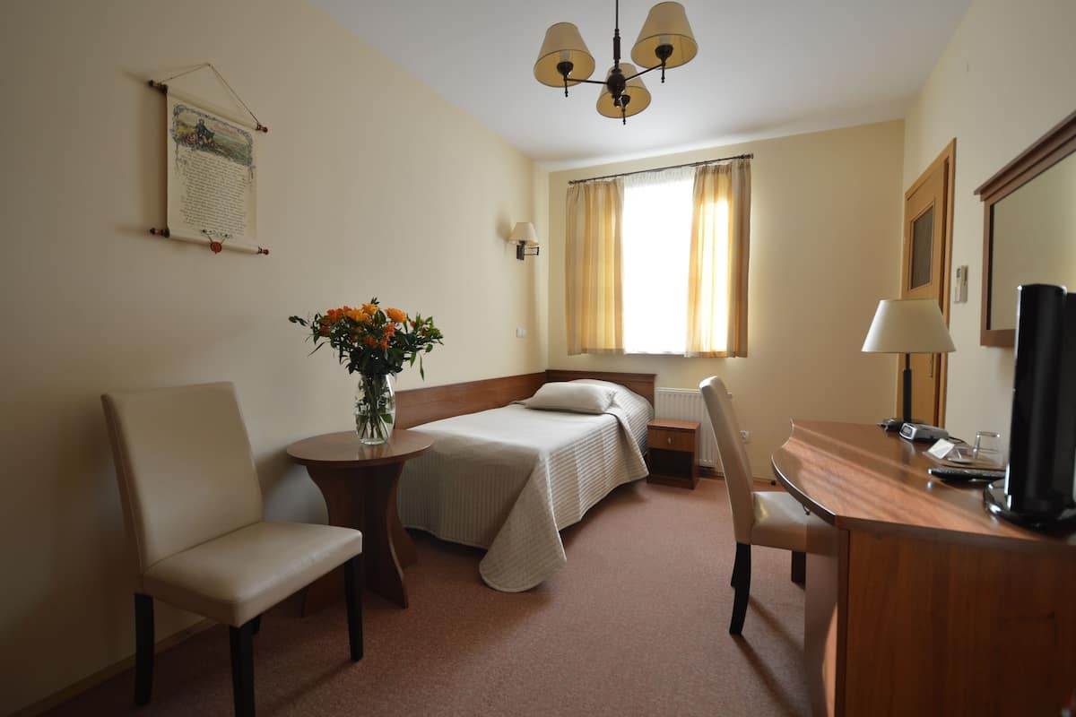 A hotel room with a bed, a desk, a chair, a small table with a vase of flowers, a lamp, and a window with curtains.