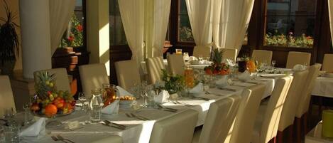 A dining area with tables set for a meal, featuring white tablecloths, chairs, and a variety of table settings including plates, glasses, and a fruit bowl.