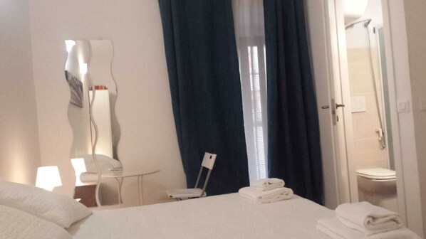Habitación de hotel con cama, escritorio, silla, espejo, ventana con cortinas y zona de ducha.