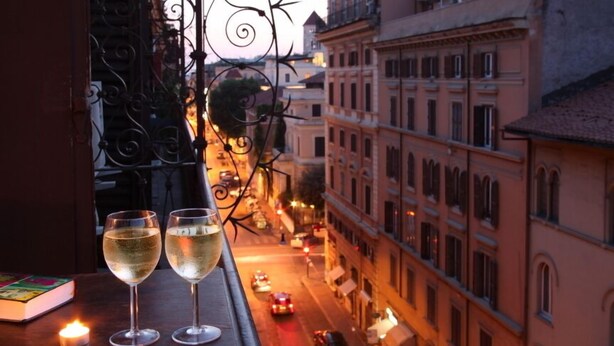 Vista desde una ventana con dos copas de vino blanco sobre una mesa, una vela y un libro.
