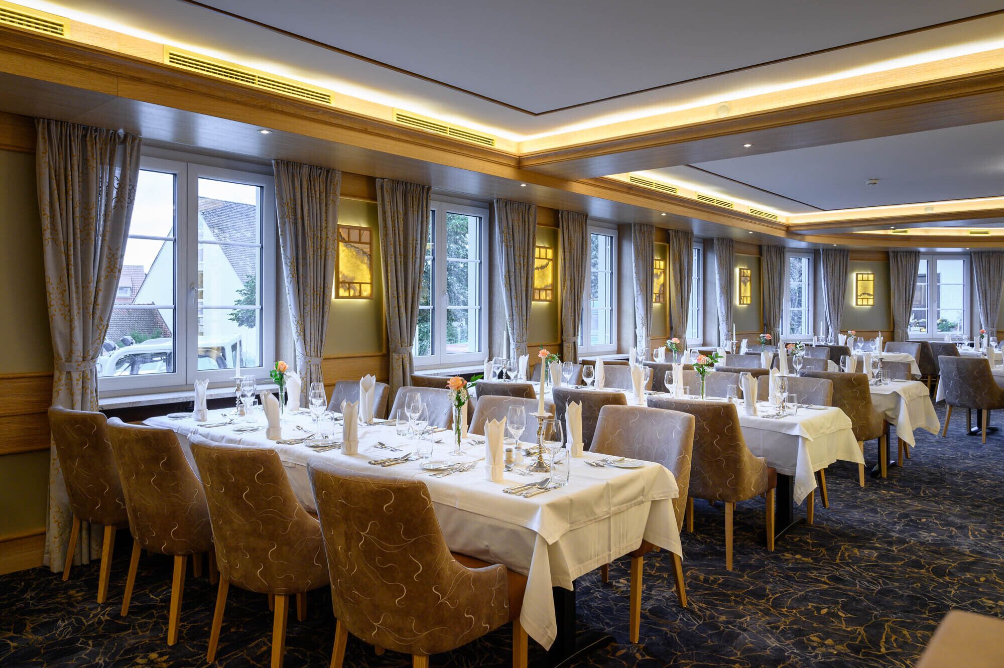 Un comedor de hotel con varias mesas dispuestas para comer, ventanas amplias con cortinas y un suelo de mármol.