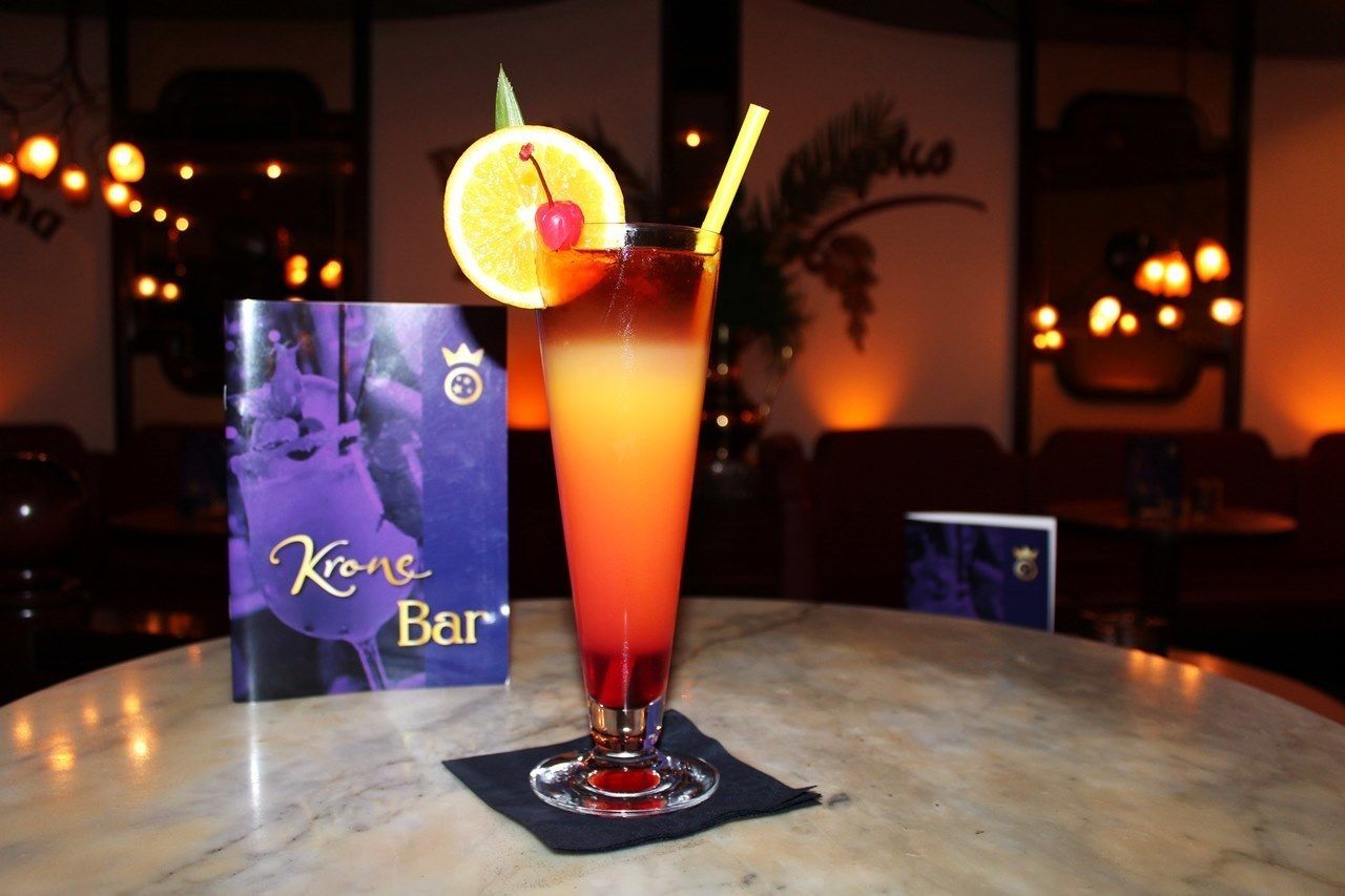 A cocktail with a cherry and orange slice garnish on a bar table, with a 'Krone Bar' sign in the background.