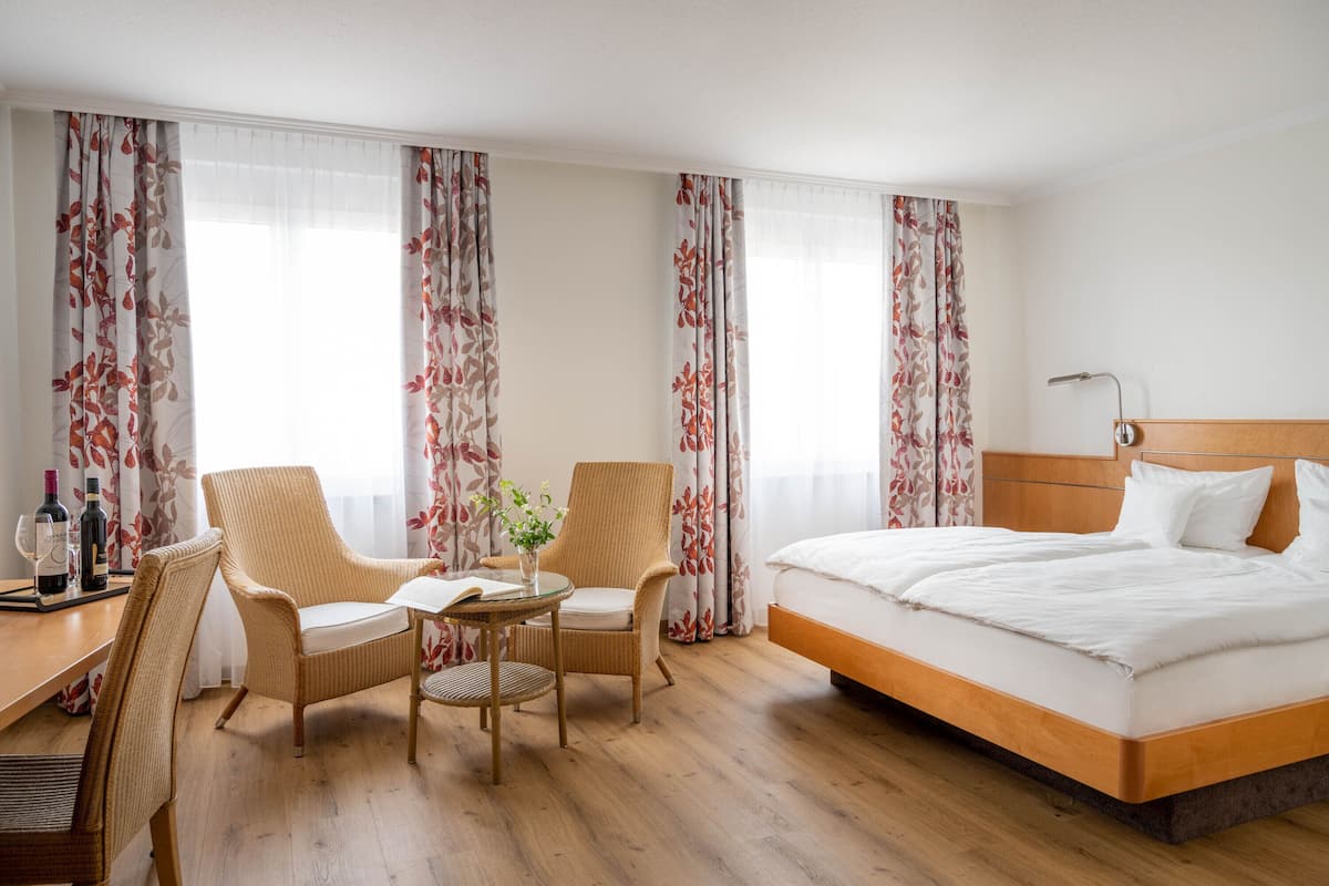 Una habitación de hotel con una cama grande, dos sillones, una mesita con un florero y un escritorio con botellas.