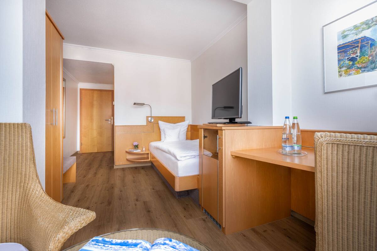 Habitación de hotel pequeña con cama, escritorio, televisor y una silla de mimbre.