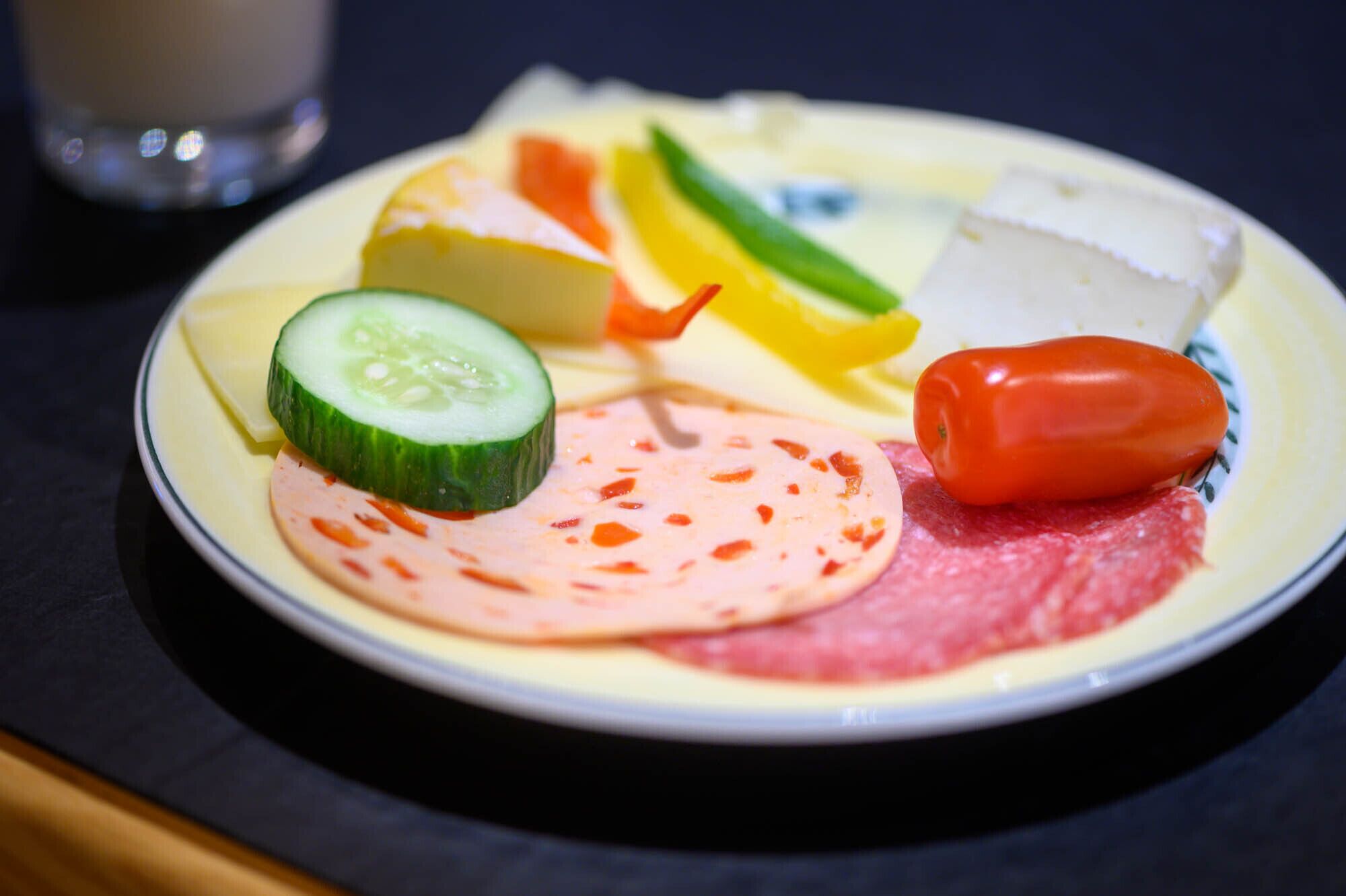 Un plato con diversos alimentos, incluyendo pepino en rodajas, queso, jamón y un pimiento rojo.