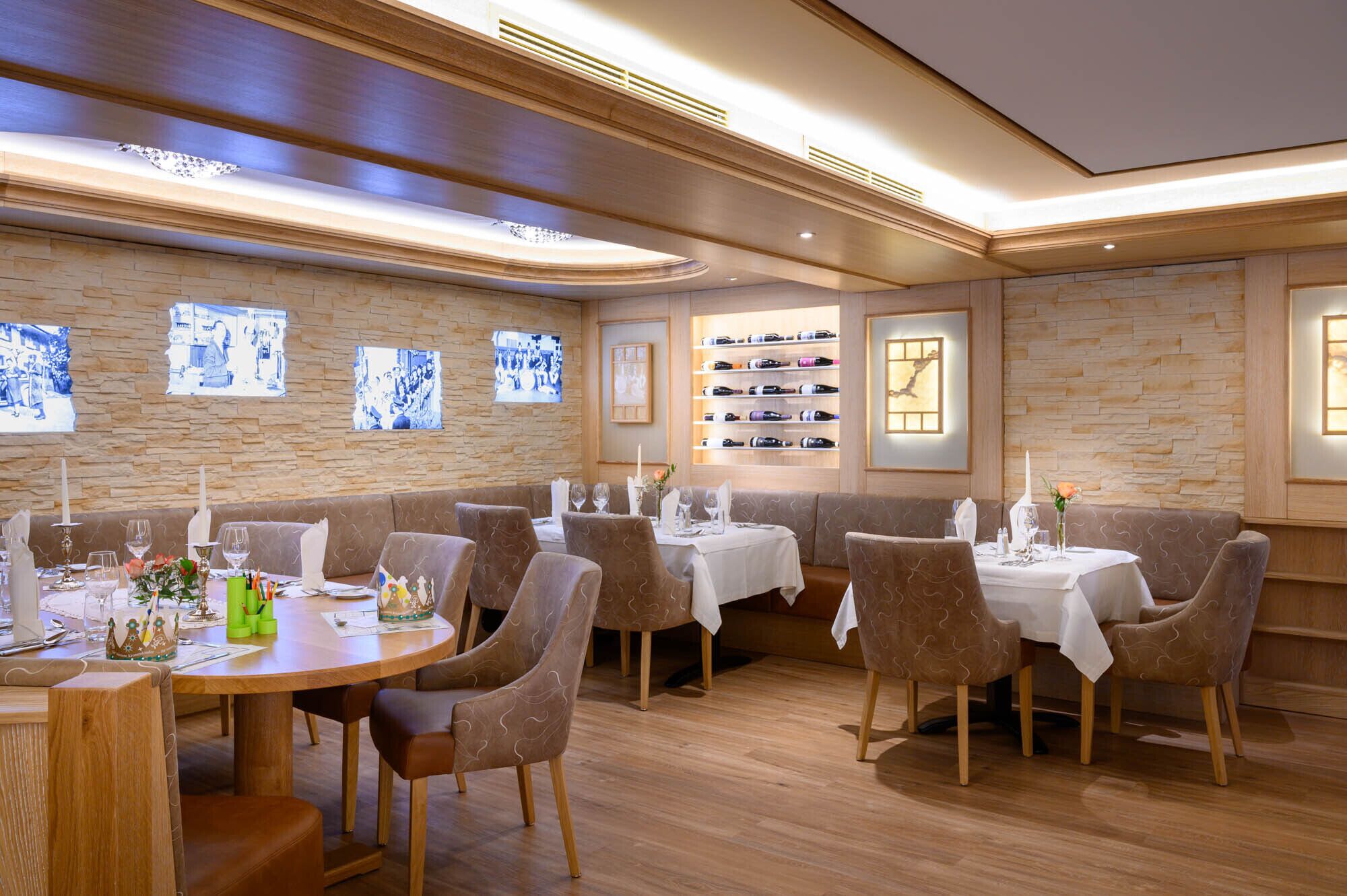 A restaurant with wooden walls, stone accents, and a wine display. Tables are set with white tablecloths and brown chairs.