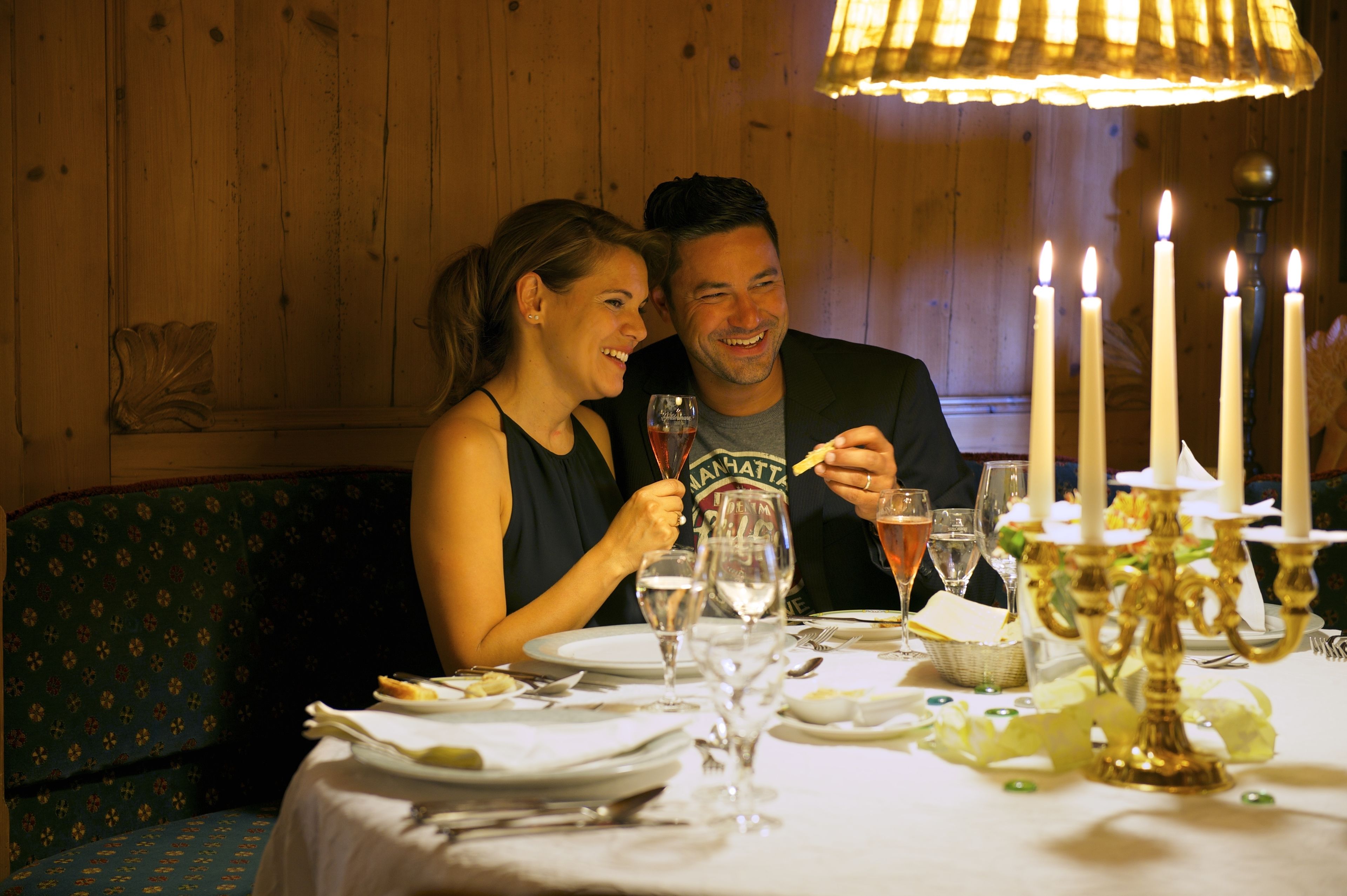 A couple dining at a table with candles and a candelabra, in a room with wooden walls and a patterned chair.